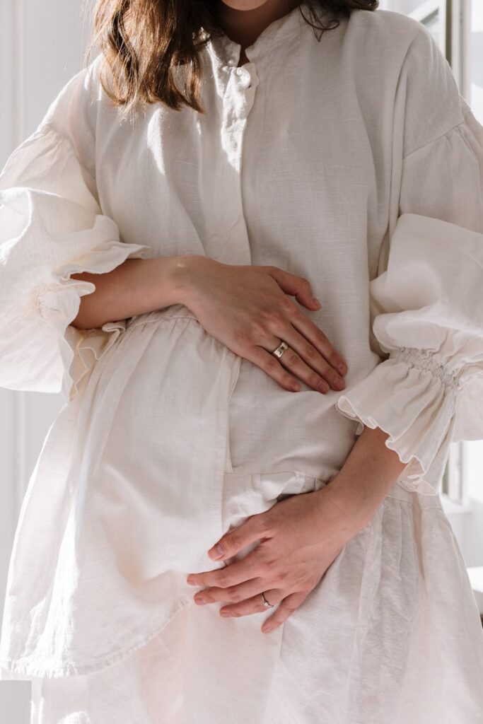 A serene image of a pregnant woman wearing a white dress, embracing her baby bump with gentle hands.